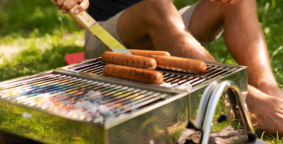 Neljä makkaraa grillataan hiiltymällä kompaktilla retkigrillillä ulkona auringonpaisteen ja vihreän ruohon loisteessa.
