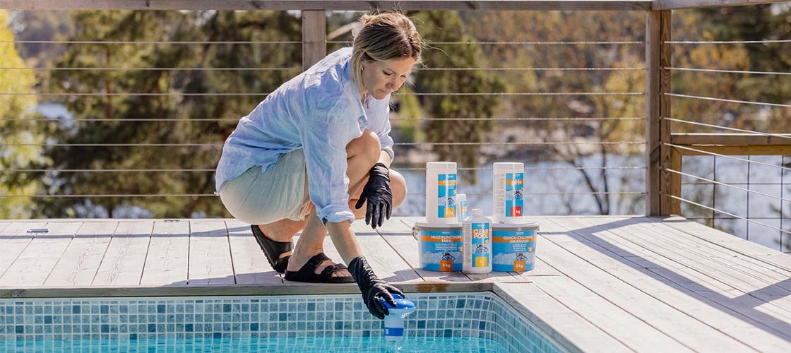 En person hukar vid kanten av en pool på en solbelyst träterrass och placerar en blå doseringsenhet i vattnet, med flera förpackningar poolkemikalier uppradade bredvid.
