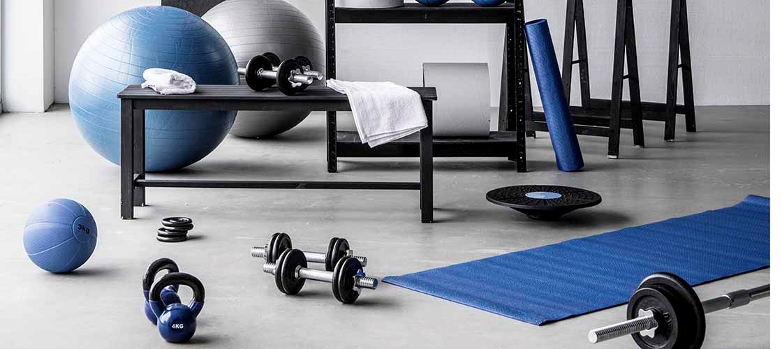 A fitness room with blue and white exercise equipment, including a yoga mat, dumbbells, a kettlebell, medicine balls, and large stability balls. A black shelving unit holds additional items such as weights, towels, and resistance bands. A hand-drawn chart labeled “Squat Challenge” is visible on the wall.