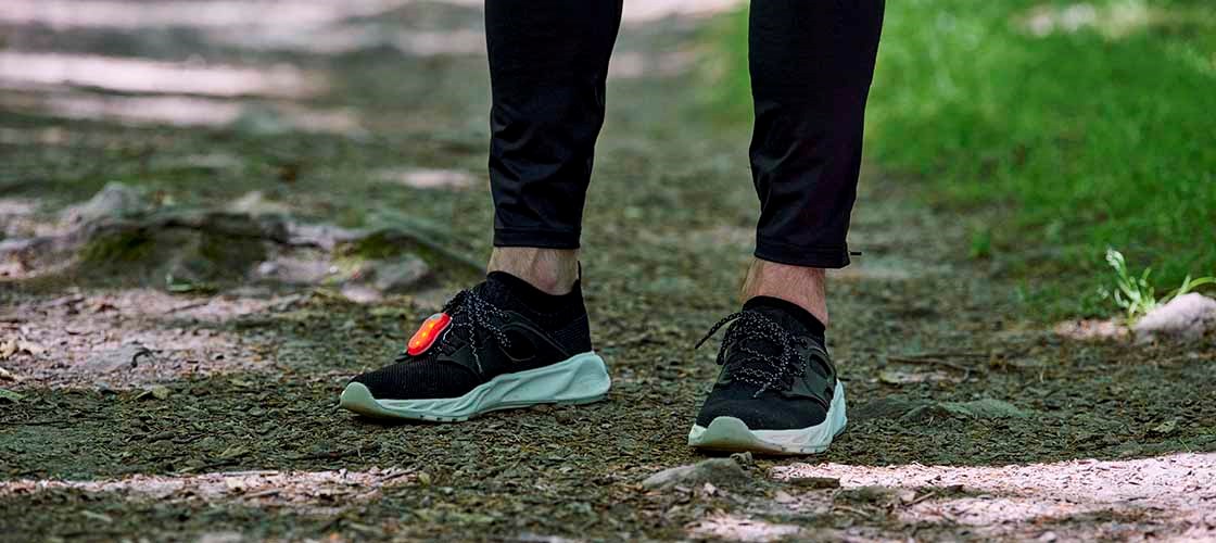 A close-up of a pair of black running shoes standing on a gravel path, with grass in the background. A small light is attached to the shoes' laces and is shining.